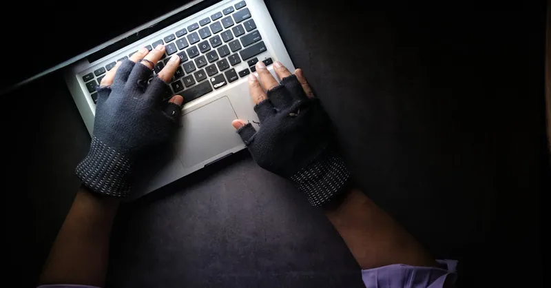 Hacker in fingerless gloves typing on laptop keyboard from above in a dark setting.