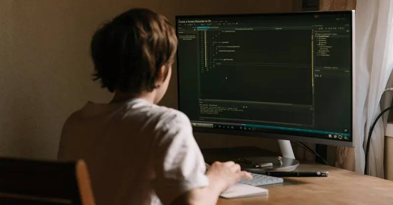 A teenager focused on coding software on a desktop monitor in a home office setting.