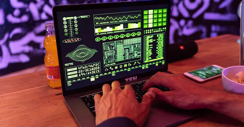 A person typing on a laptop displaying green code with an orange bottle and smartphone nearby.